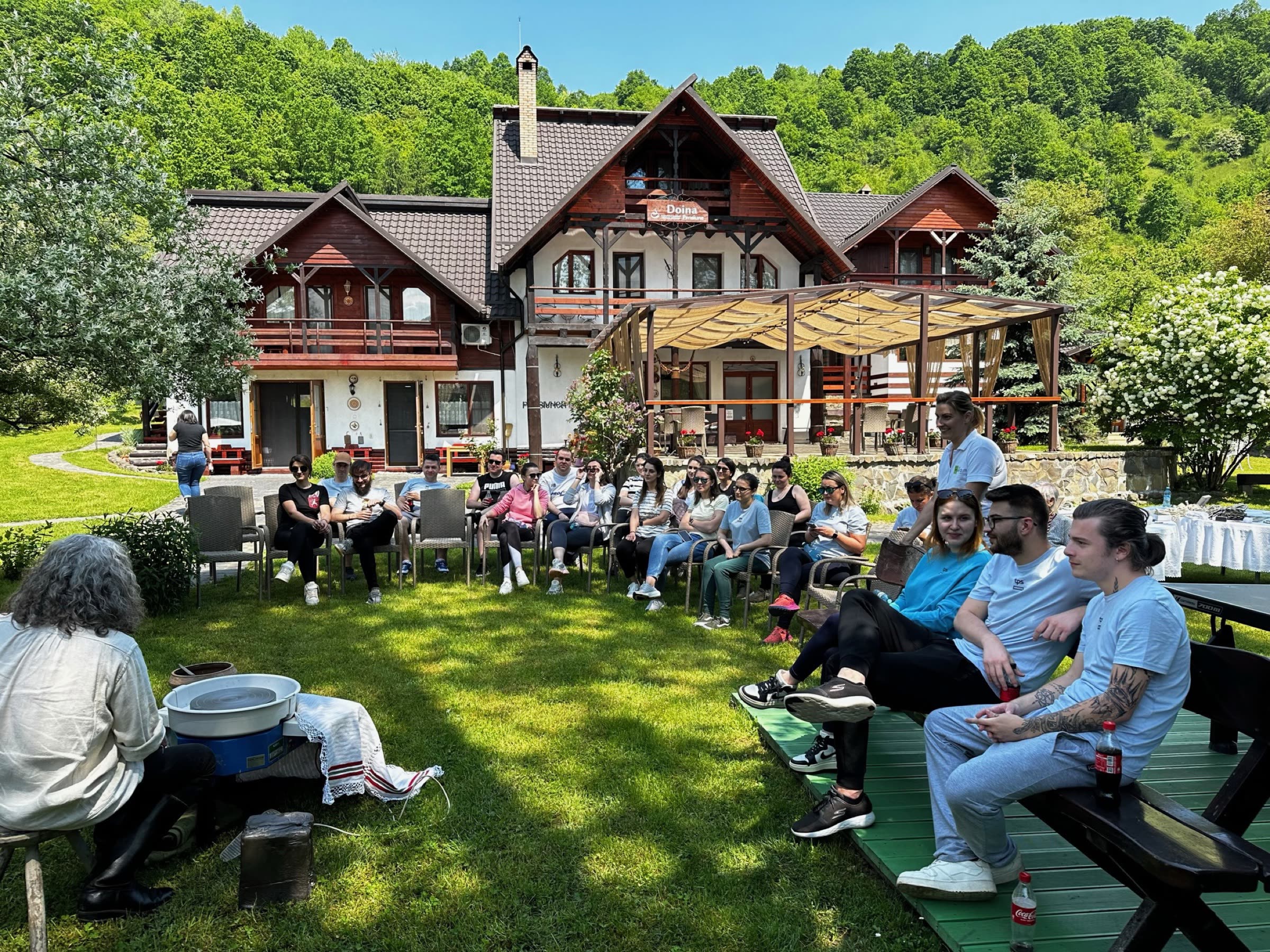 Grup mare, așezat în semicerc pe iarbă în fața pensiunii, la o zi de team building