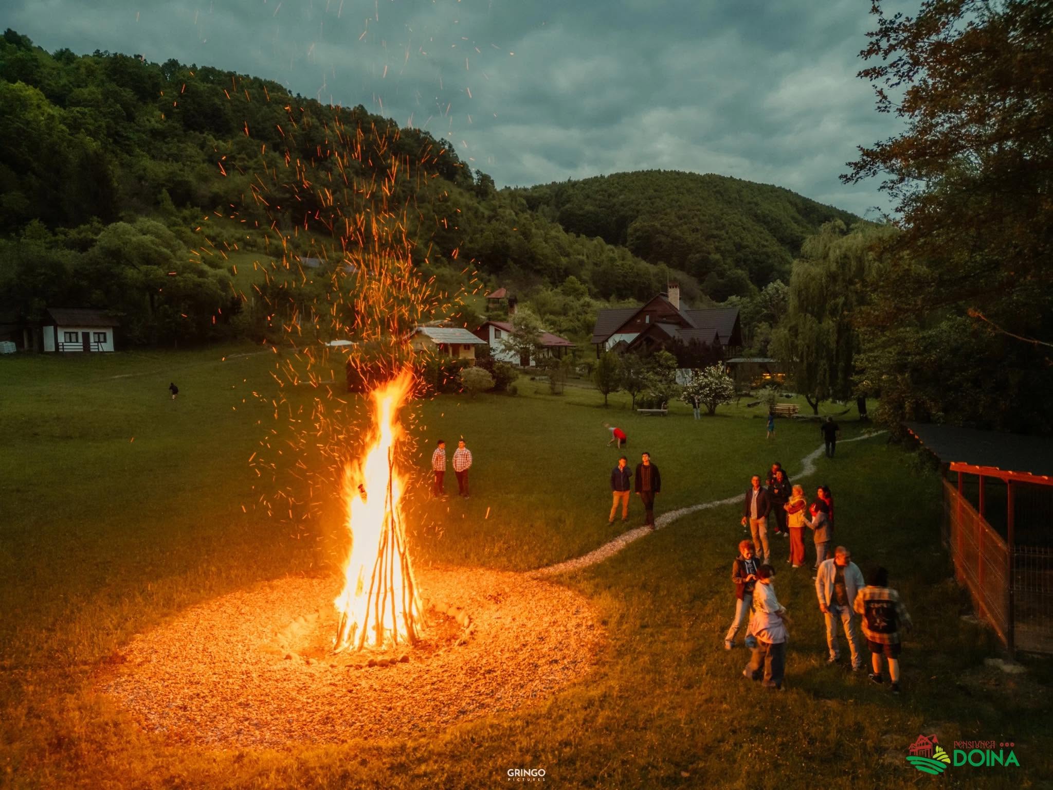Foc de tabără mare în grădina domeniului, cu oaspeții adunați, la asfințit